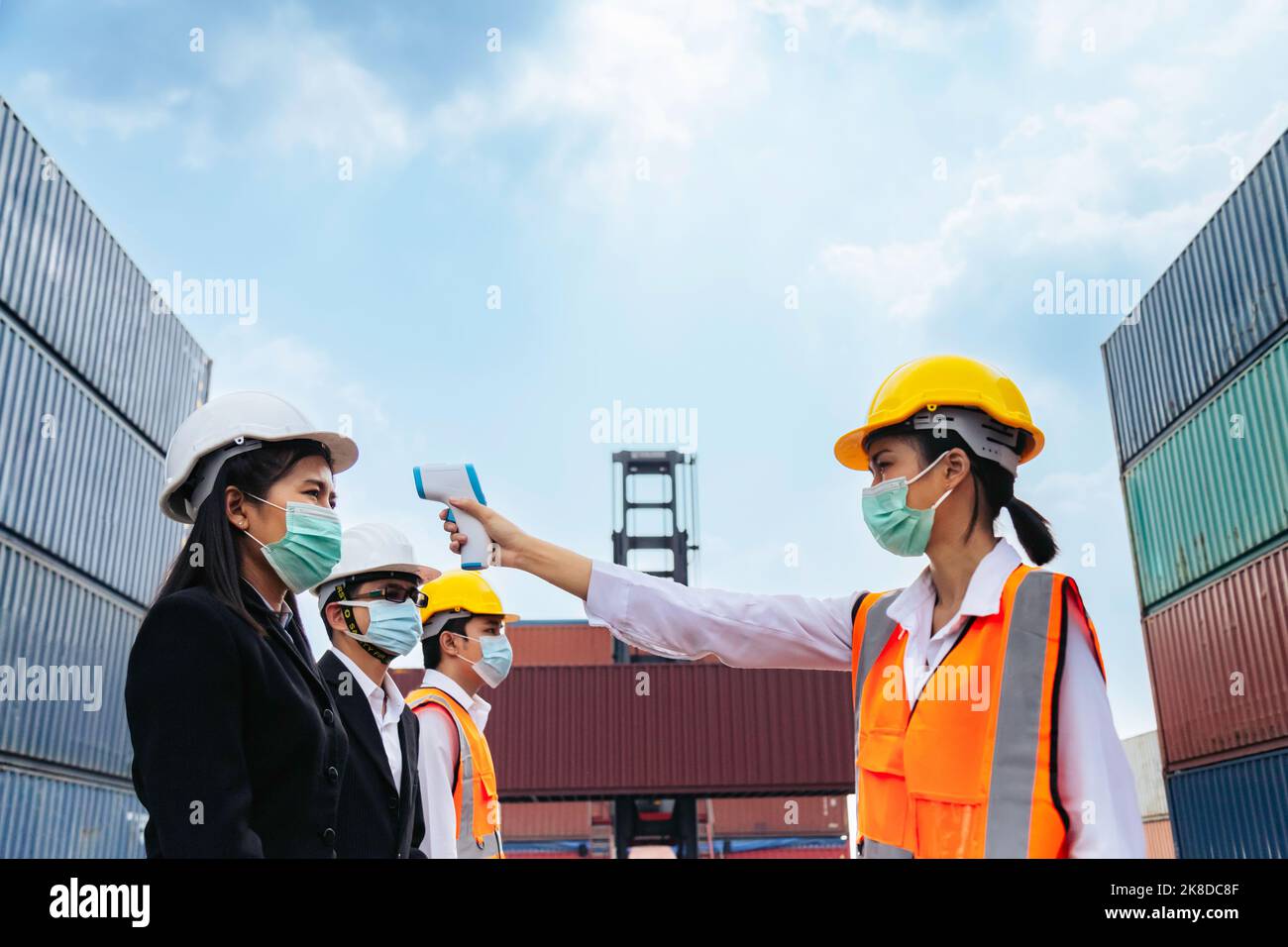 asiatische Geschäftsfrau und Team tragen Gesichtsmaske Überprüfung Fieber durch Digitalthermometer vor dem Betreten der Arbeit in logistischen Fracht-Lager bei Manufacturin Stockfoto