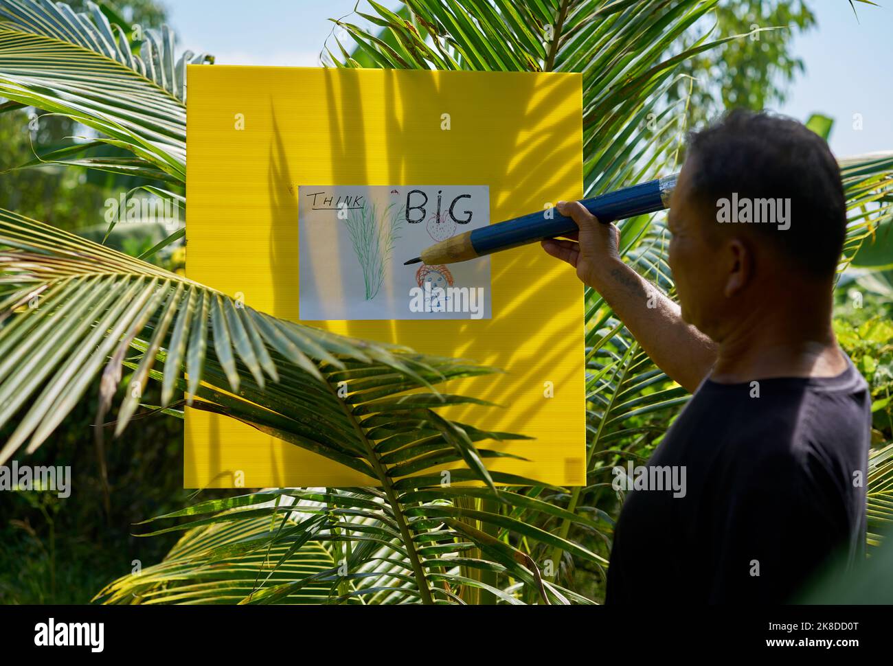 Ein Künstler, der in einem tropischen Garten mit einem sehr großen Bleistift zeichnet. Stockfoto