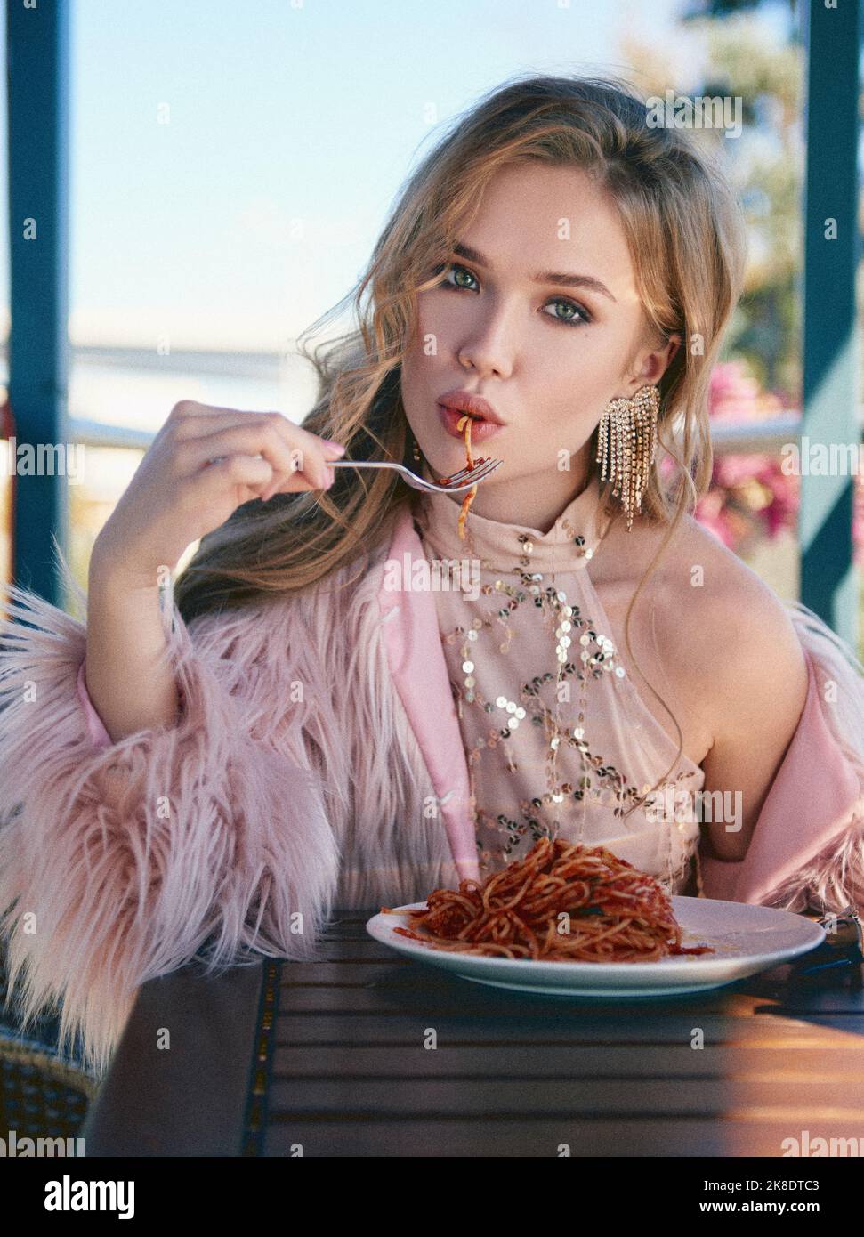 Hübsches Mädchen, das in einem Café sitzt und Pasta (Spaghetti) isst. Porträt einer stilvollen jungen Frau im Restaurant, die Kleid und Pelzmantel trägt. Film Korn effe Stockfoto