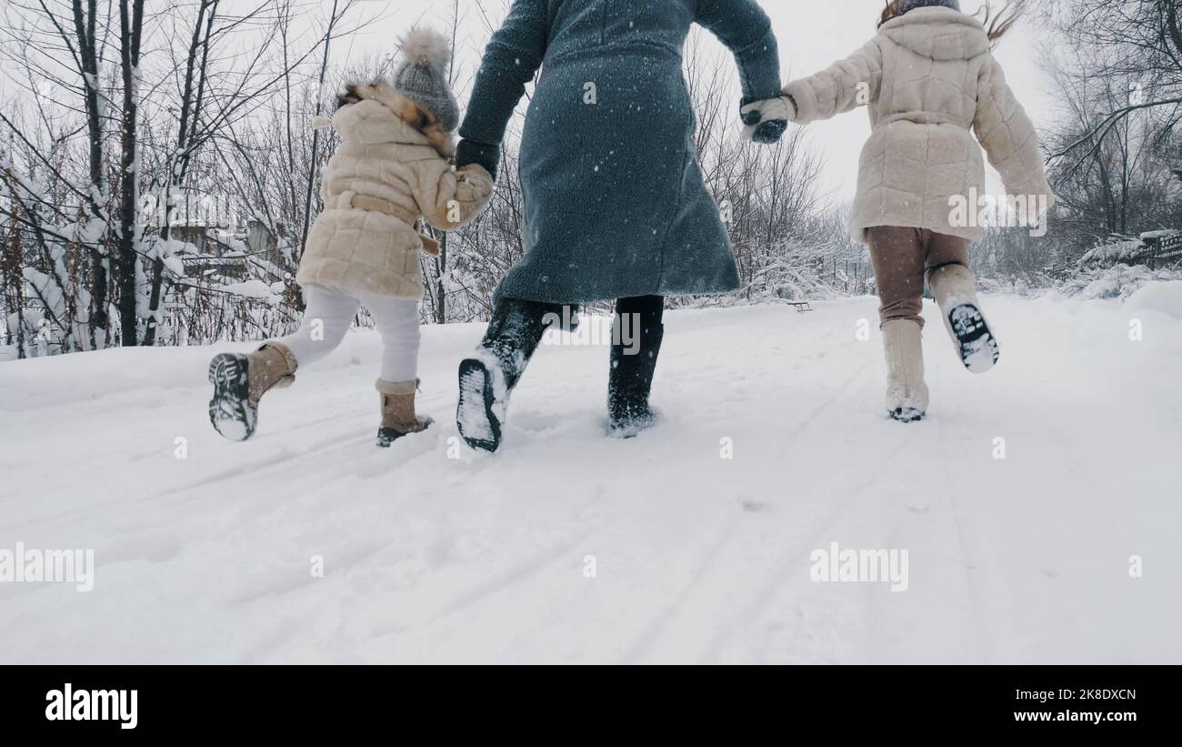 Winter Familienaktivitäten im Freien. Rückansicht. Glückliche Frau mit 2 Töchtern laufen auf verschneiten Straßen, im Wald, bei Schneefall. Familie hat Spaß, Zeit zusammen am verschneiten Wintertag zu verbringen. Zeitlupe. Hochwertige Fotos Stockfoto