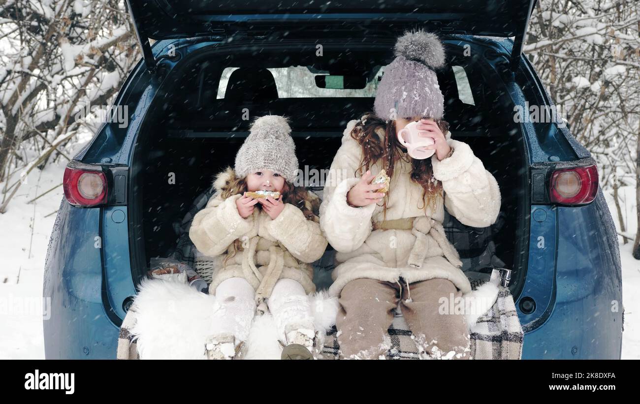 Winter-Tee-Picknick. Happy Cutie, kleines Mädchen, in warmen Winterkleidung gekleidet, haben leckere Snacks, Tee-Party im Freien. Sie sitzen auf dem Kofferraum, im verschneiten Wald, bei Schneefall. Winterspaß für die ganze Familie. Glückliche Zeit am verschneiten Wintertag. Winteraktivitäten für die ganze Familie im Freien. Hochwertige Fotos Stockfoto