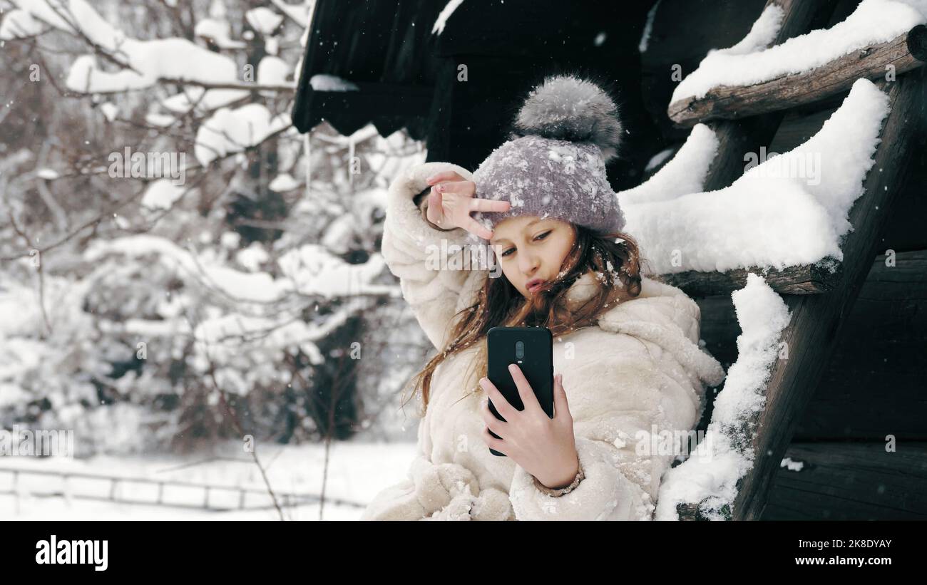 Verschneite Winter-Selfie. Cutie-Mädchen, gekleidet in warme Winterkleidung, voll mit Schneeflocken bedeckt, sitzt auf der Bank und nimmt Selfie, mit dem Smartphone, im Freien, während Schneefall, in der Nähe von alten Holzhaus. Verschneiten Wintertag. Hochwertige Fotos Stockfoto