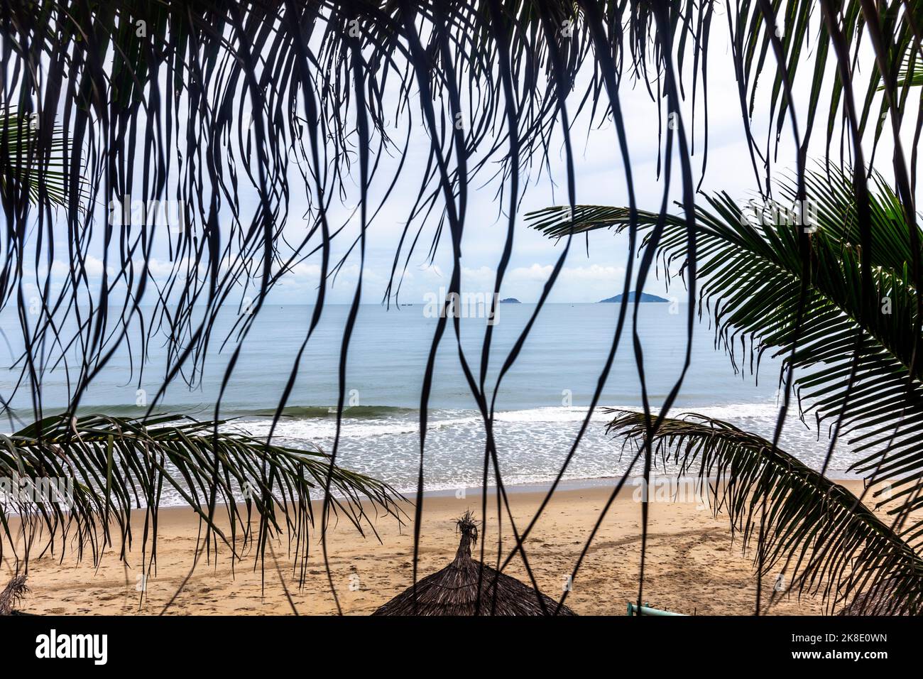Ein erhöhter Blick auf das Meer mit Strandhütten und Fußabdrücken im Sand, die durch ein kleines Fenster mit Palmen blicken. Stockfoto