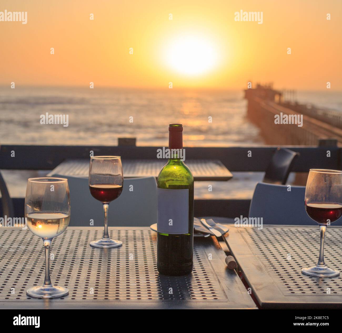 Flasche Rotwein mit drei Gläsern auf einem Tisch, mit einem wunderschönen Hintergrund bei Sonnenuntergang - Swakopmund Pier, Namibia Stockfoto