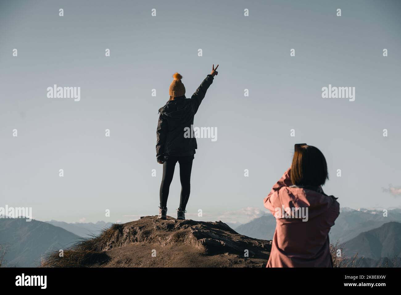 kaukasisches Mädchen in Kapuzenjacke braun Beanie und schwarze Hosen stehen auf dem Rücken auf Stein mit Arm ausgestreckt und Finger bilden einen Buchstaben V als sie Stockfoto