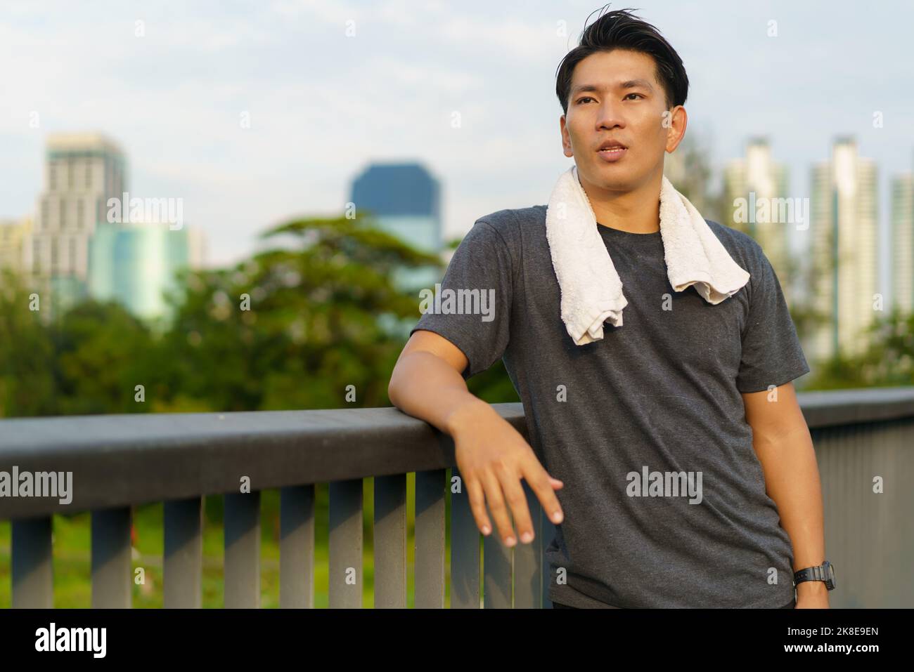Porträt eines jungen asiatischen Mannes, der im Freien auf einem Gehweg mit Stadtgebäude im Stehen und Schweiß abwischt und Sportkleidung trägt. Stockfoto