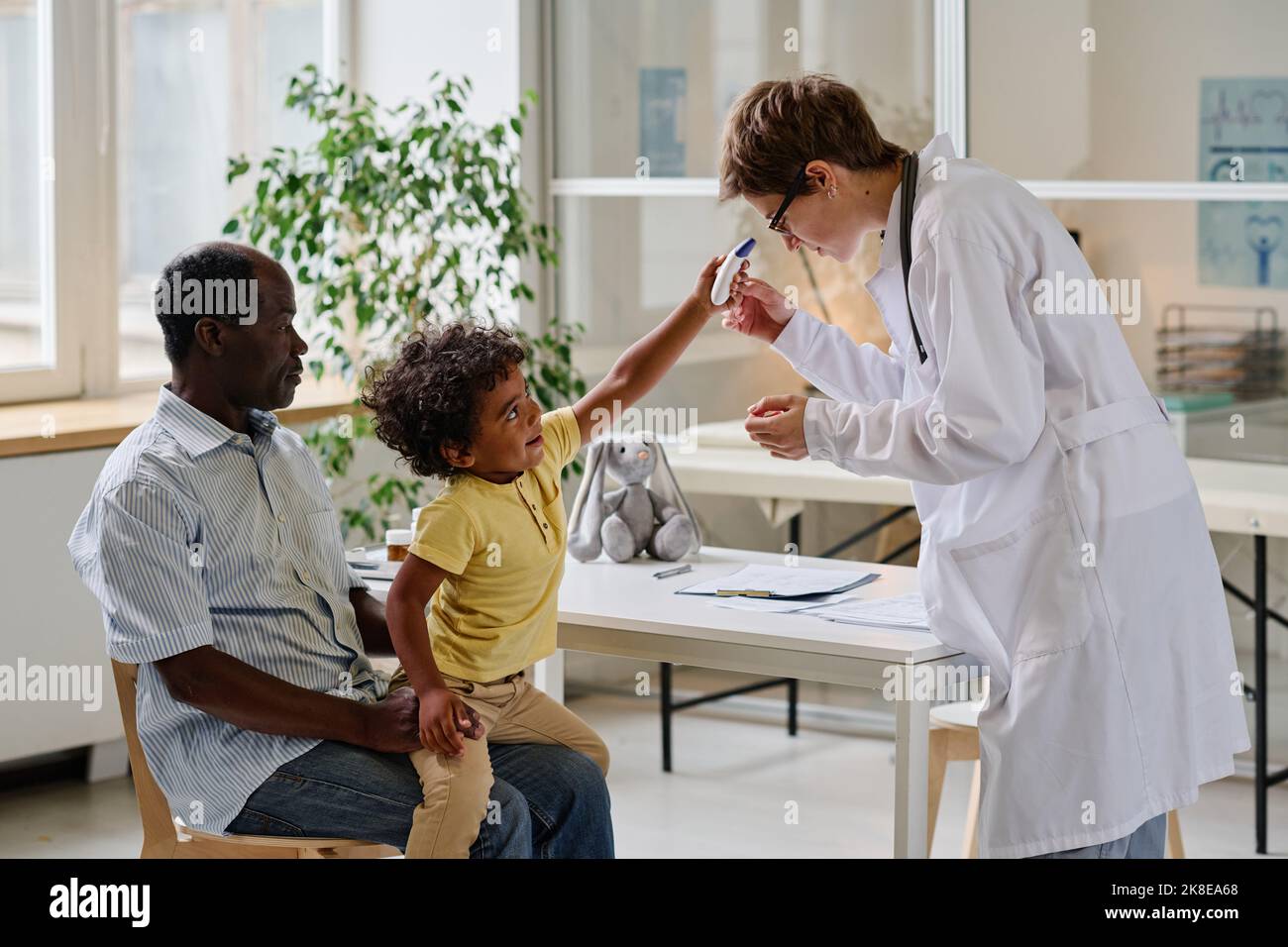 Ärztin, die versucht, das Kind mit speziellen medizinischen Geräten zu untersuchen, während er mit seinem Vater in der Klinik sitzt Stockfoto