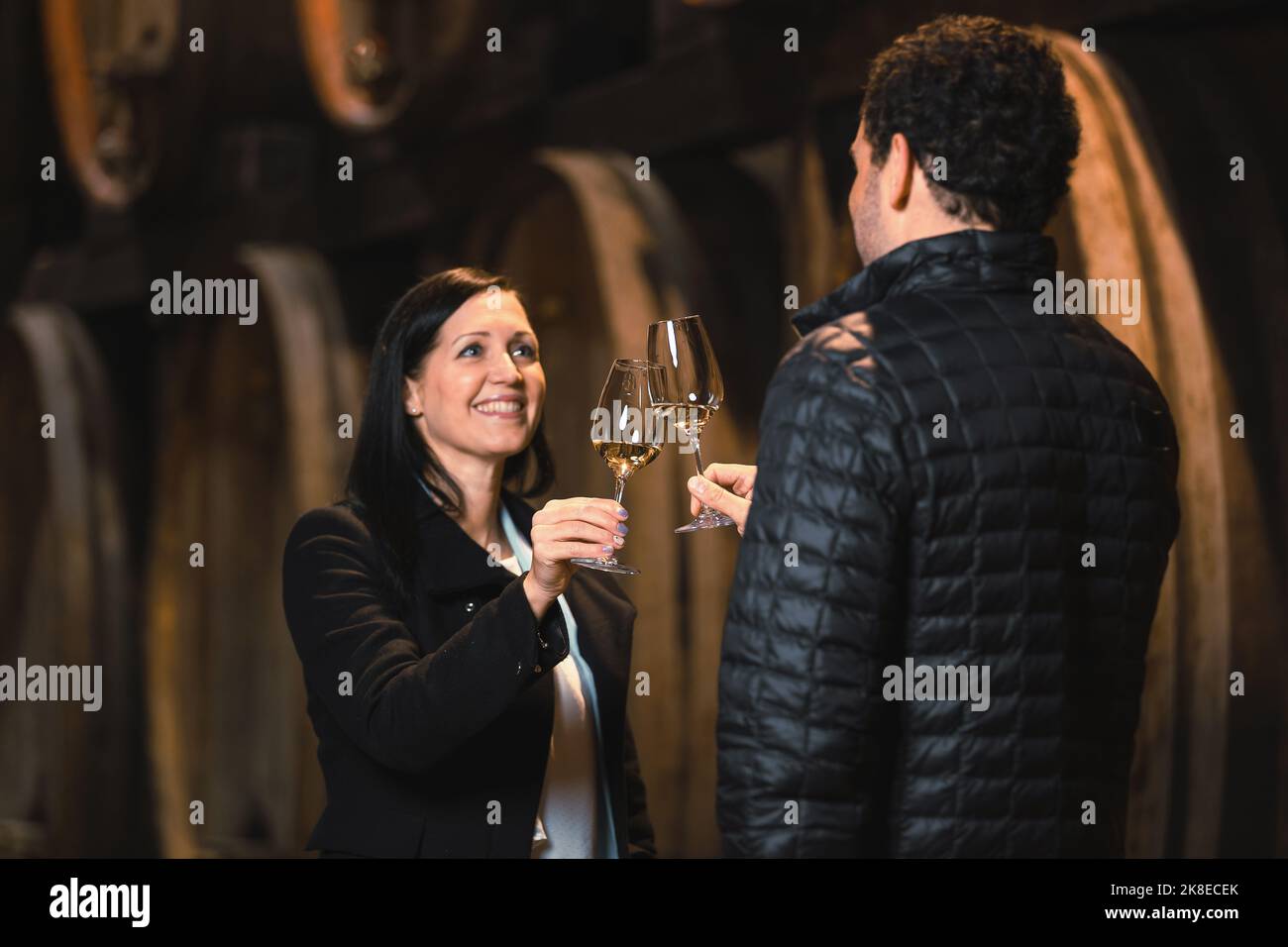 Mittlere Ansicht des kaukasischen Paares während einer Weinprobe in einem Weinkeller, mit großen Eichenfässern im Hintergrund Stockfoto