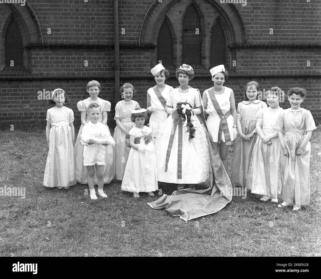 1962, historisch, Maifeiertag, draußen auf dem Gelände einer Kirche und der diesjährigen Maikönigin, stehend mit anderen Kindern in ihren langen Kleidern für das traditionelle Fest, das den Sommer feiert, Leeds, England, Großbritannien. Ein kleiner Junge, der einzige Mann, der das Kissen für die Krone hält. Stockfoto