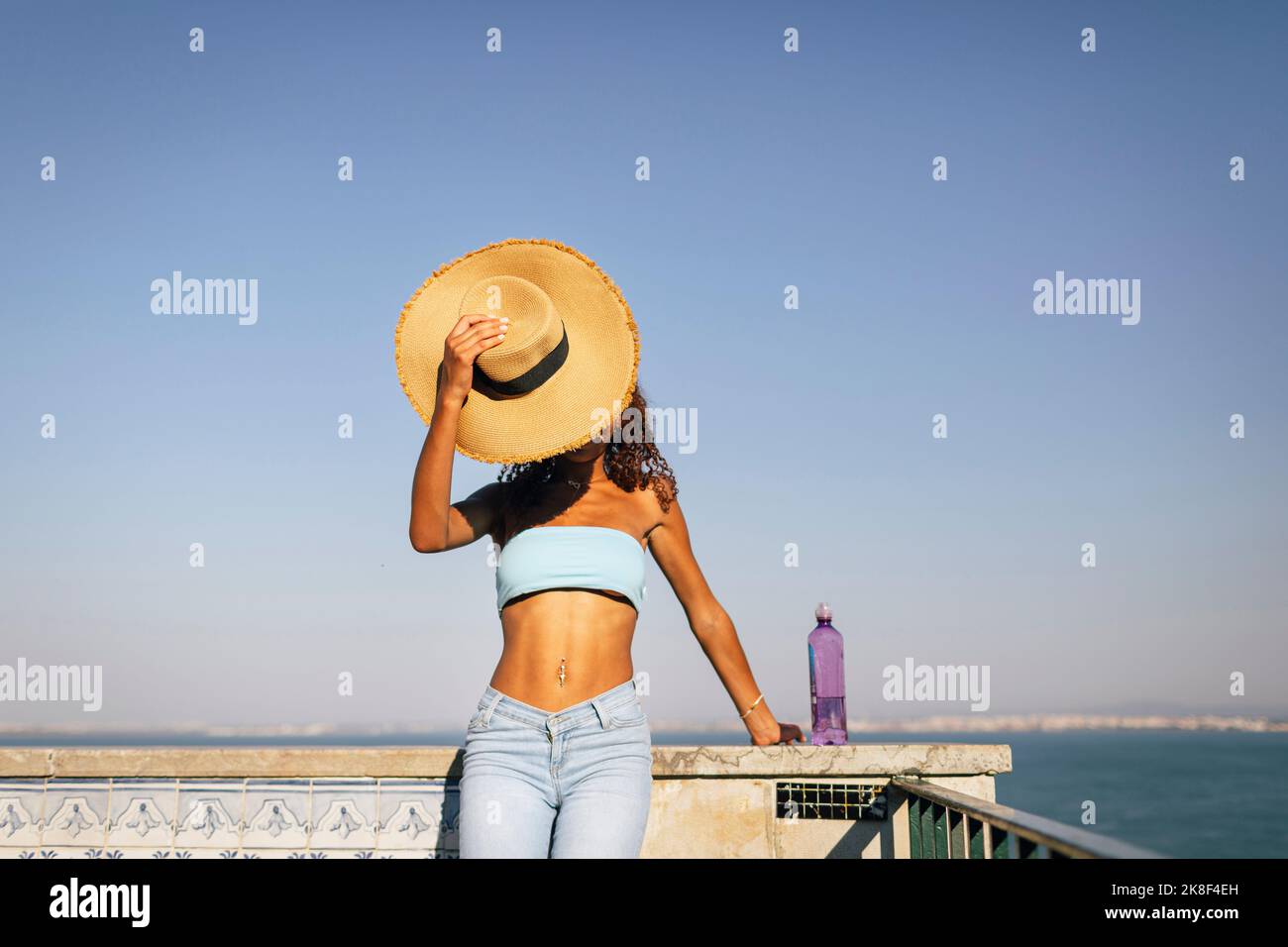 Junge Frau bedeckt das Gesicht mit einem Hut, der an einem sonnigen Tag an der Wand steht Stockfoto