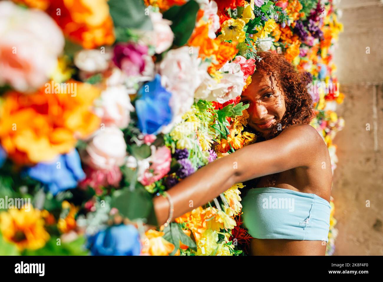 Lächelnde junge Frau, die sich an einer mehrfarbigen Blumenwand lehnt Stockfoto