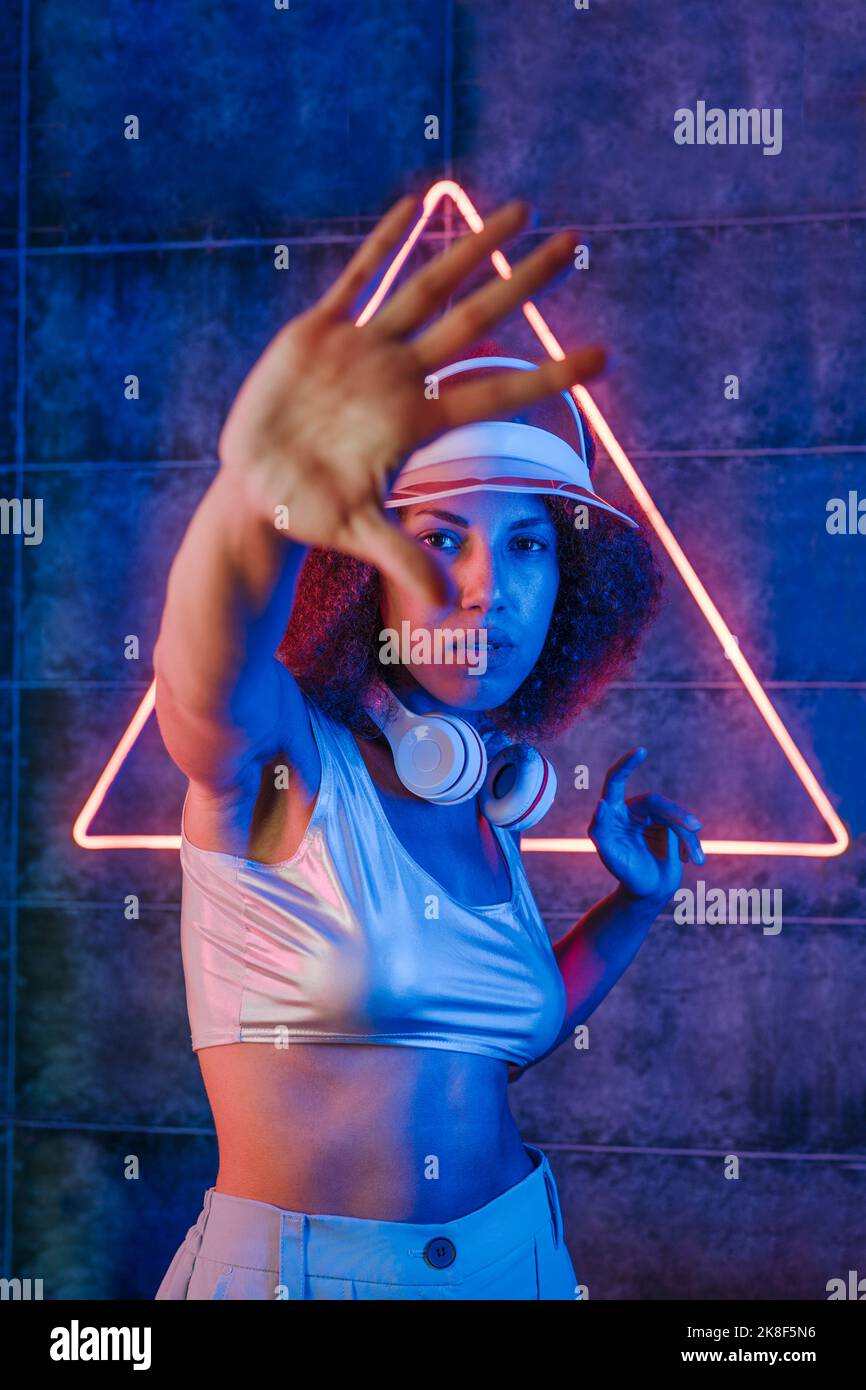 Frau mit kabellosen Kopfhörern macht eine Stoppgeste vor dem dreieckigen Neonlicht an der Wand Stockfoto