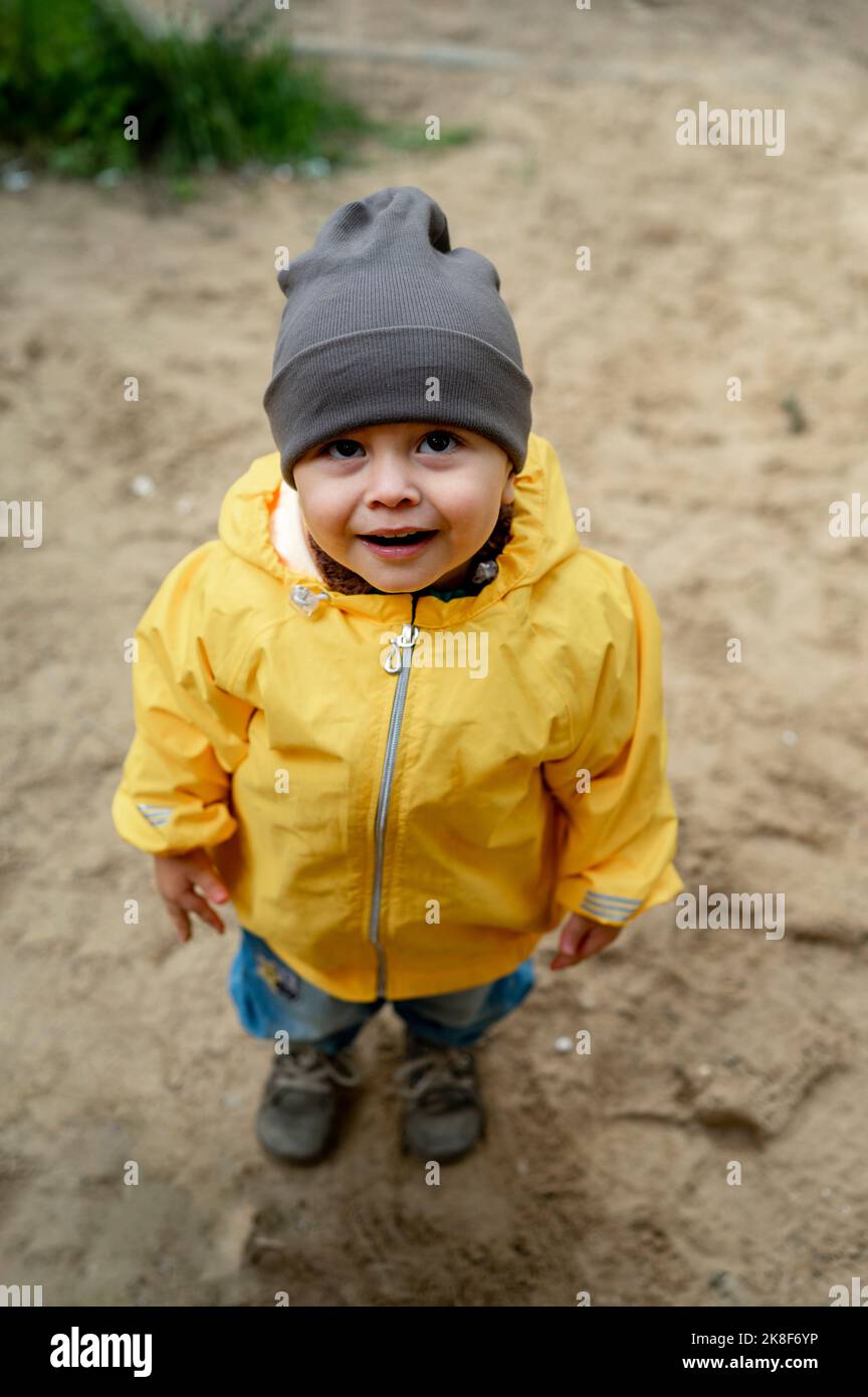 Niedlicher Baby-Junge mit Strickmütze und gelber Regenjacke auf Sand Stockfoto