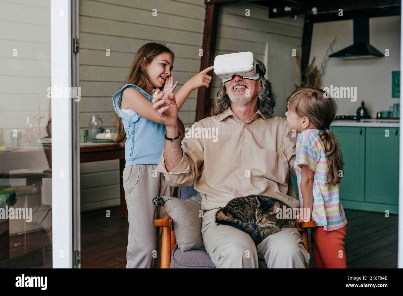 Lächelnder Großvater mit VR-Brille, die von Enkelinnen zu Hause gestikelt wird Stockfoto