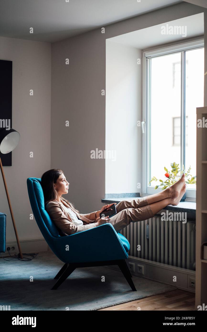 Nachdenkliche Geschäftsfrau, die zu Hause mit den Füßen auf einem Stuhl sitzt Stockfoto