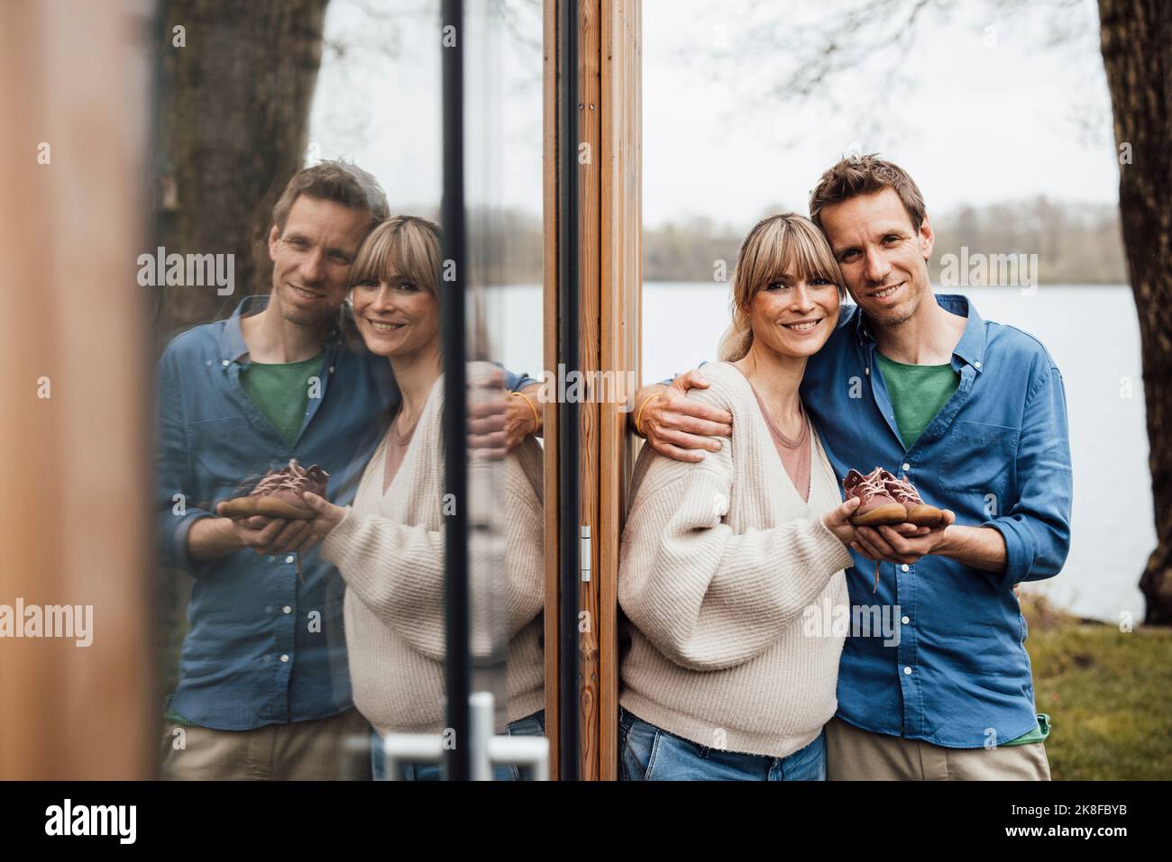 Lächelnde werdende Eltern halten Babyschühchen durch Glasreflexion Stockfoto