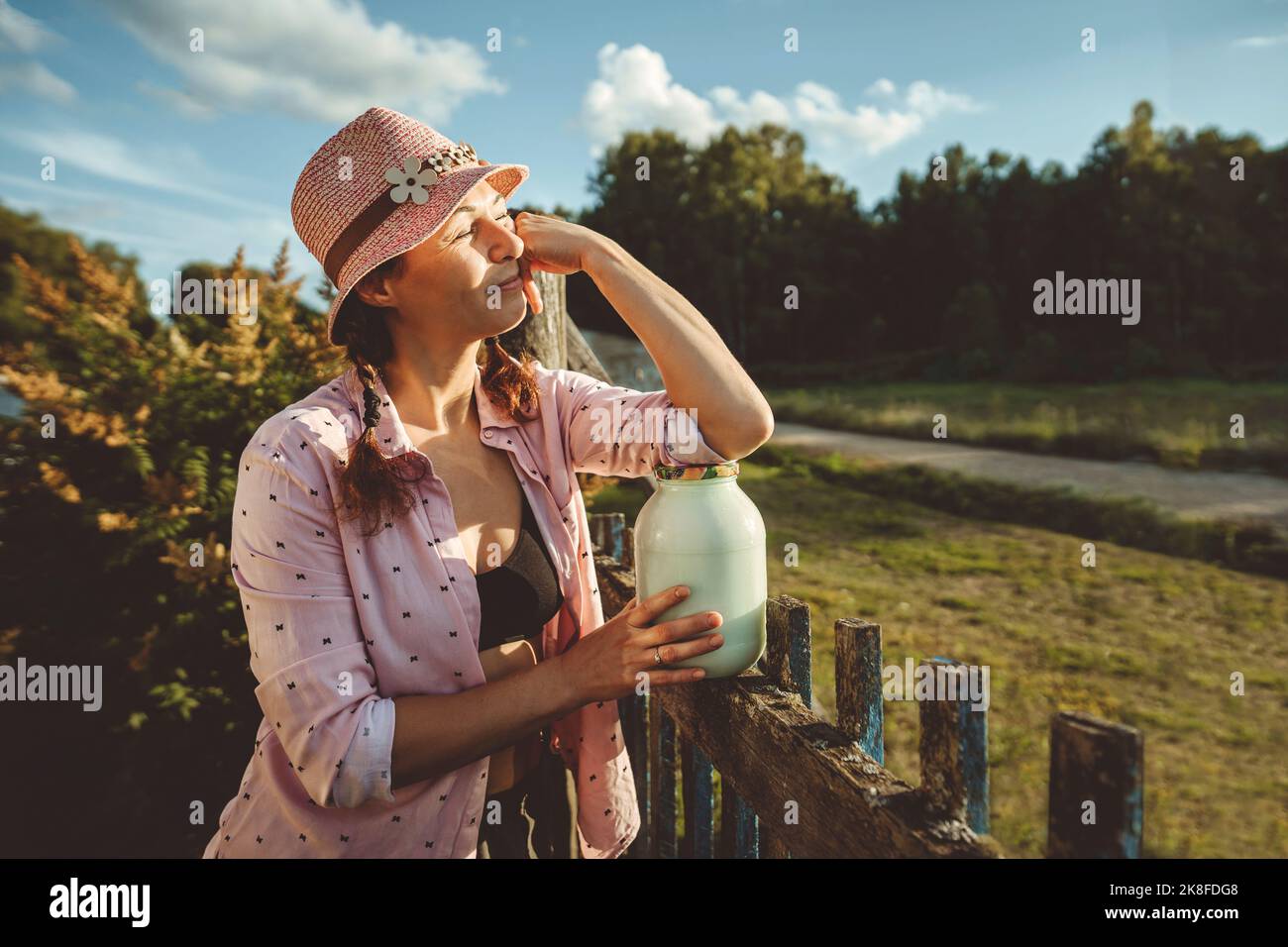 Frau, die an einem sonnigen Tag mit einem Glas voller Milch am Zaun steht Stockfoto