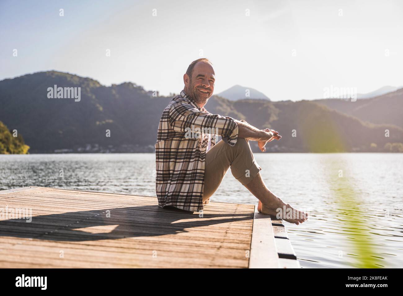 Lächelnder Mann, der im Urlaub auf dem Steg über dem See sitzt Stockfoto