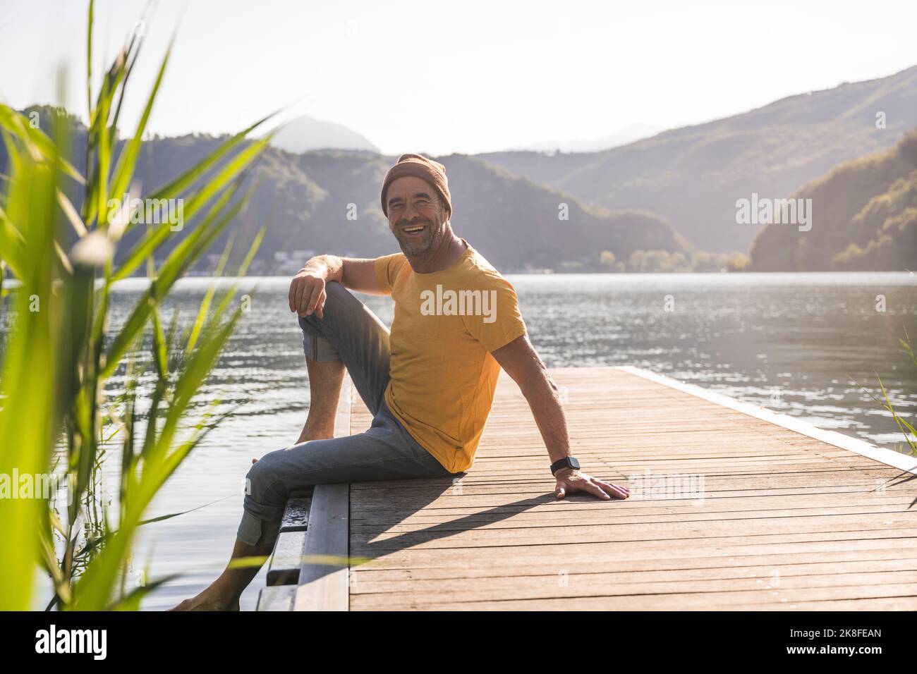 Glücklicher reifer Mann, der im Urlaub auf dem Steg über dem See sitzt Stockfoto
