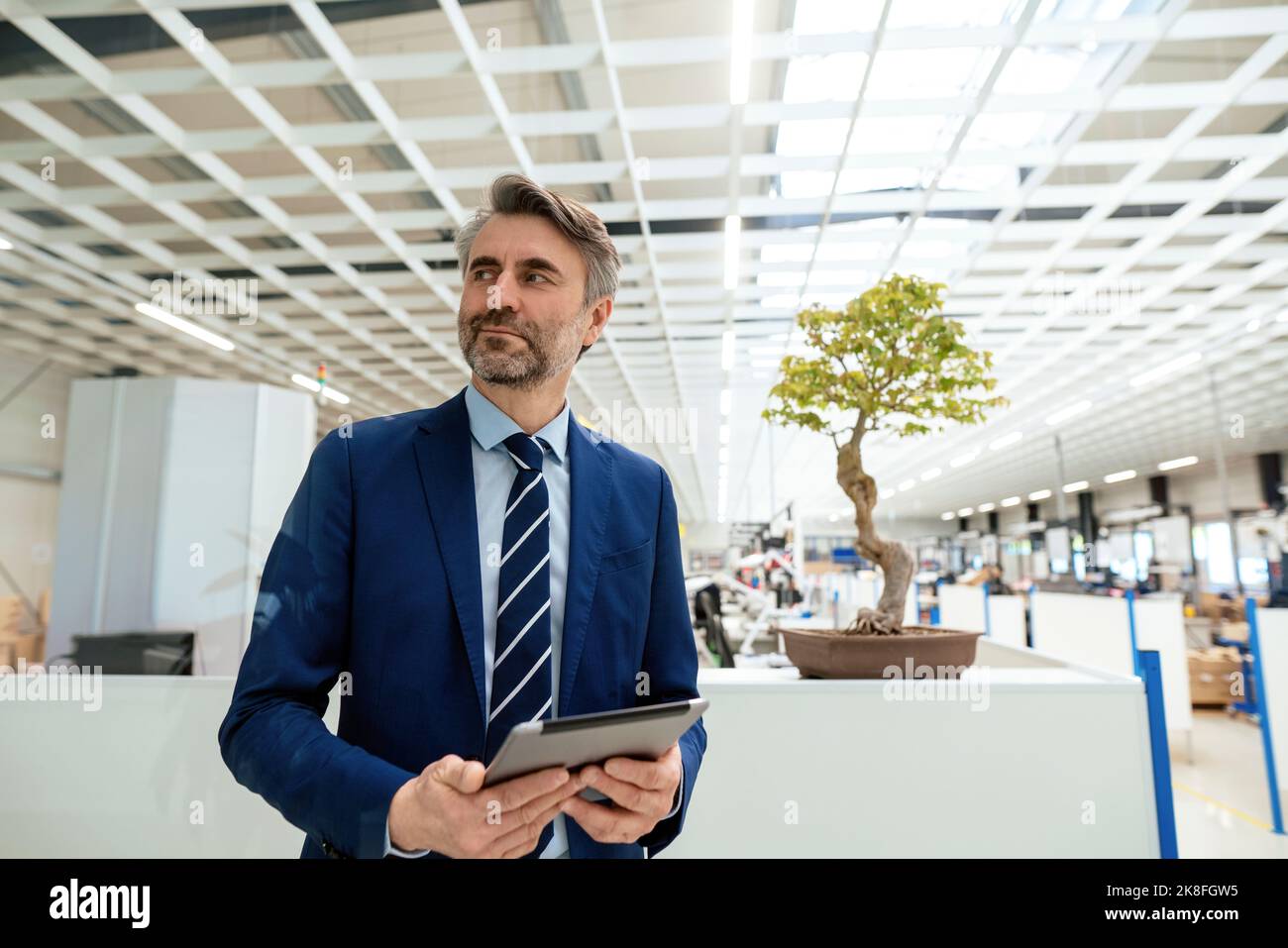 Lächelnder, reifer Geschäftsmann mit Tablet-PC, der vor dem Bonsai-Baum in der Industrie steht Stockfoto