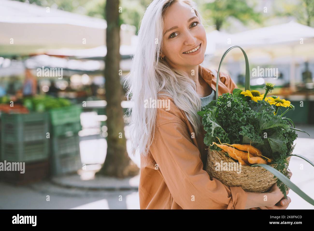 Lächelnde Frau mit Blumen und Gemüse in wiederverwendbarem Weidensack auf dem Markt Stockfoto