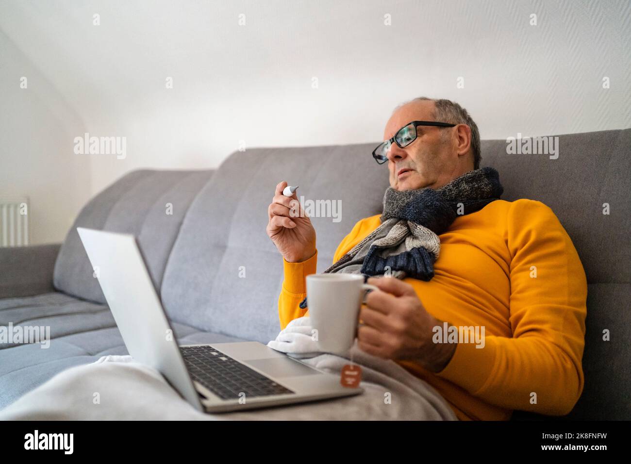 Mann mit Laptop, der den Becher hält und Fieber durch das Thermometer auf dem Sofa überprüft Stockfoto