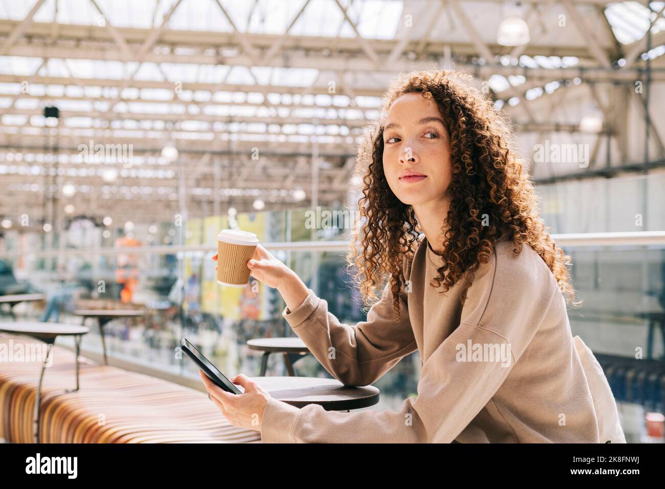 Junge Frau, die mit dem Mobiltelefon sitzt und Kaffee am Bahnhof trinkt Stockfoto