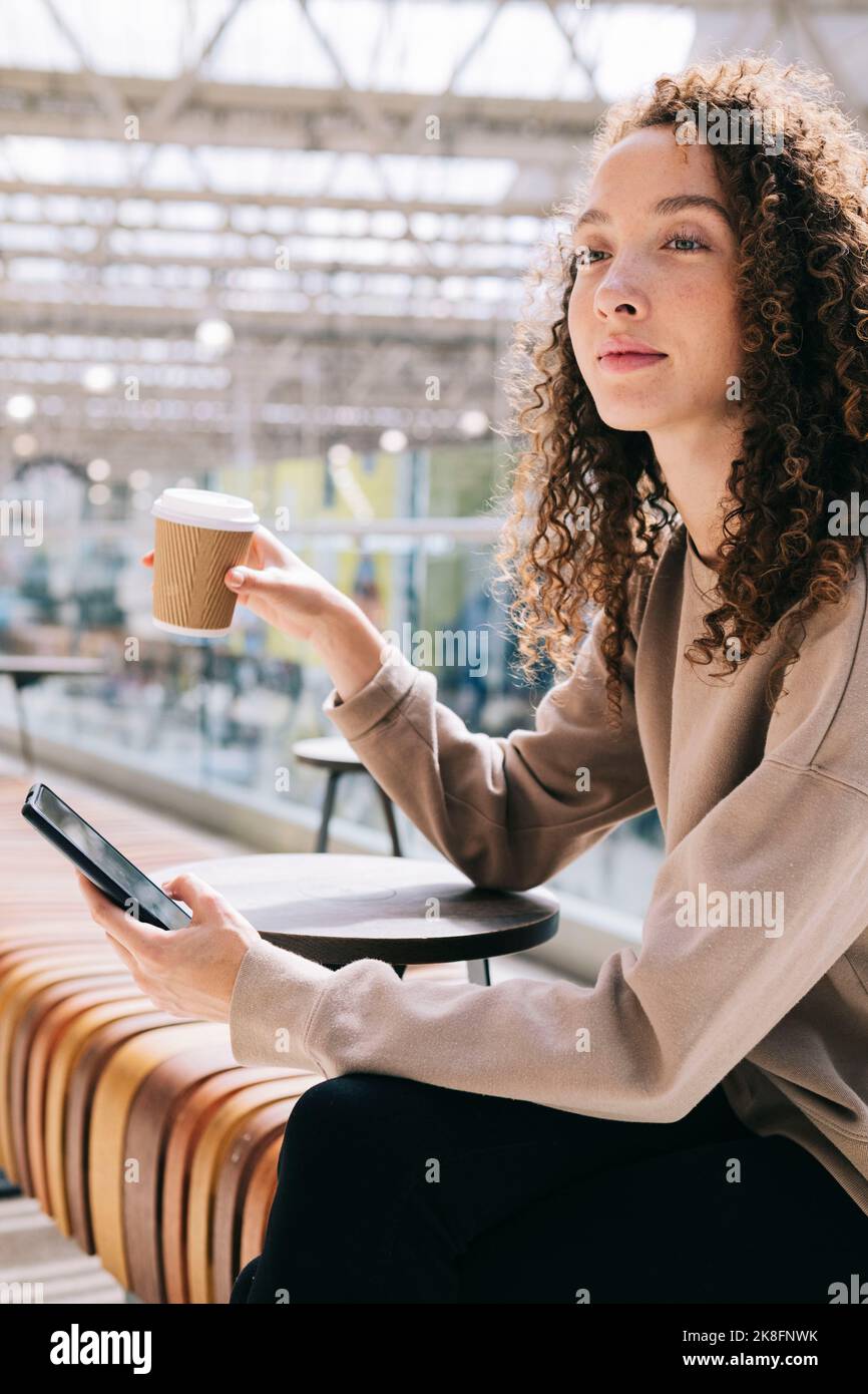 Nachdenkliche Frau, die mit dem Mobiltelefon am Bahnhof sitzt und Kaffee trinkt Stockfoto