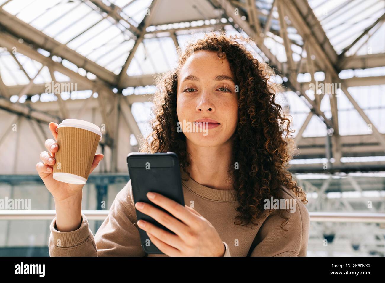Lächelnde Frau, die mit dem Mobiltelefon am Bahnhof sitzt und Kaffee trinkt Stockfoto