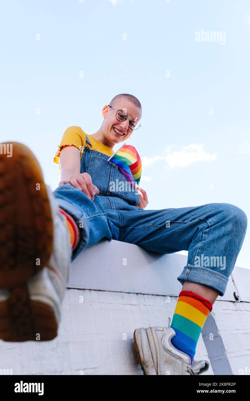 Glückliche, nicht-binäre Person mit Regenbogenfahne an der Wand Stockfoto
