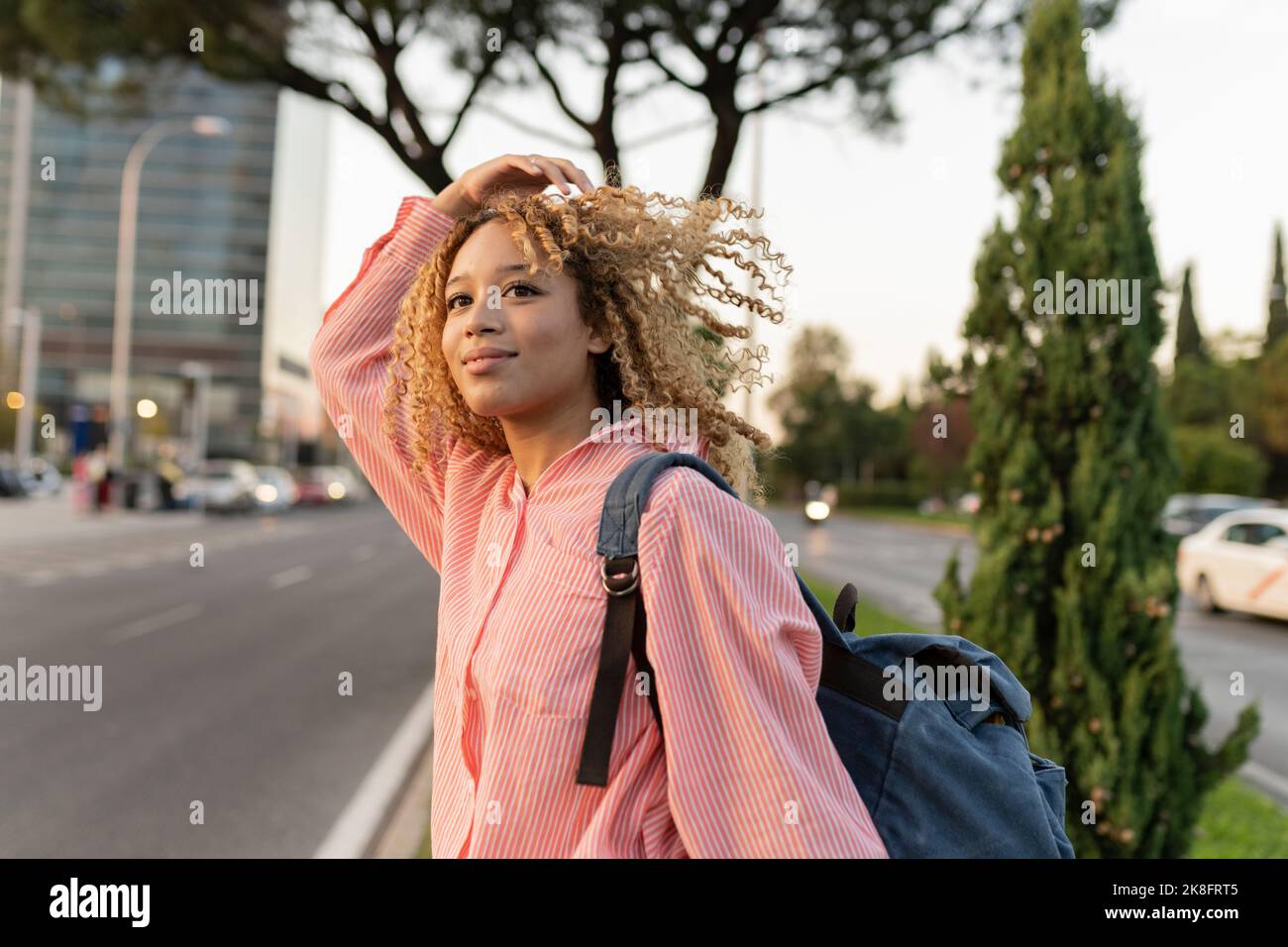 Junge Frau mit lockigen blonden Haaren trägt Rucksack überqueren Straße Stockfoto