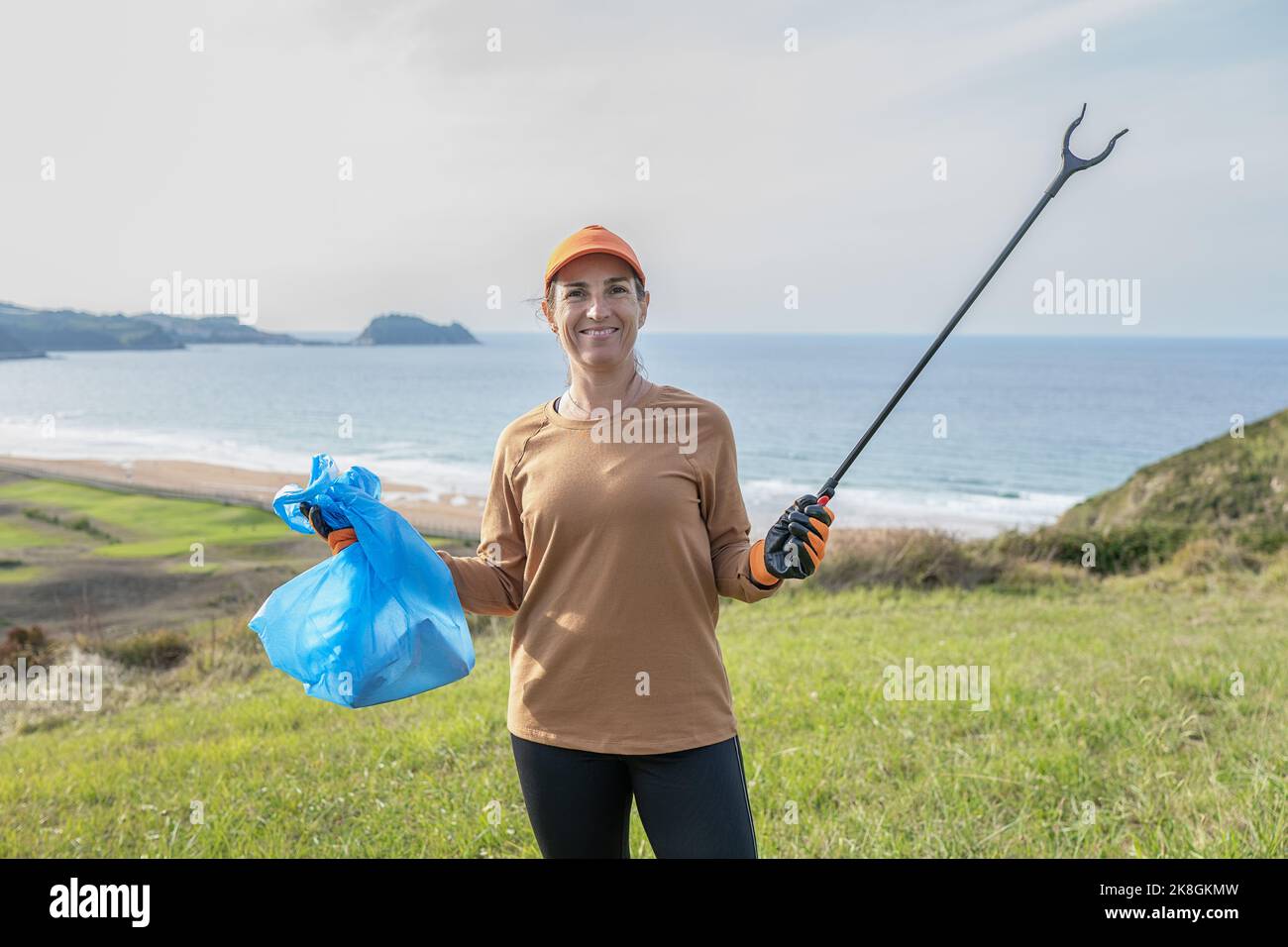 Fröhliche weibliche Freiwillige in Schutzhandschuhen und Hut in erhobenen Händen Trash Zange und Müllbeutel voll von Abfall nach Müllabfuhr auf Gras halten Stockfoto