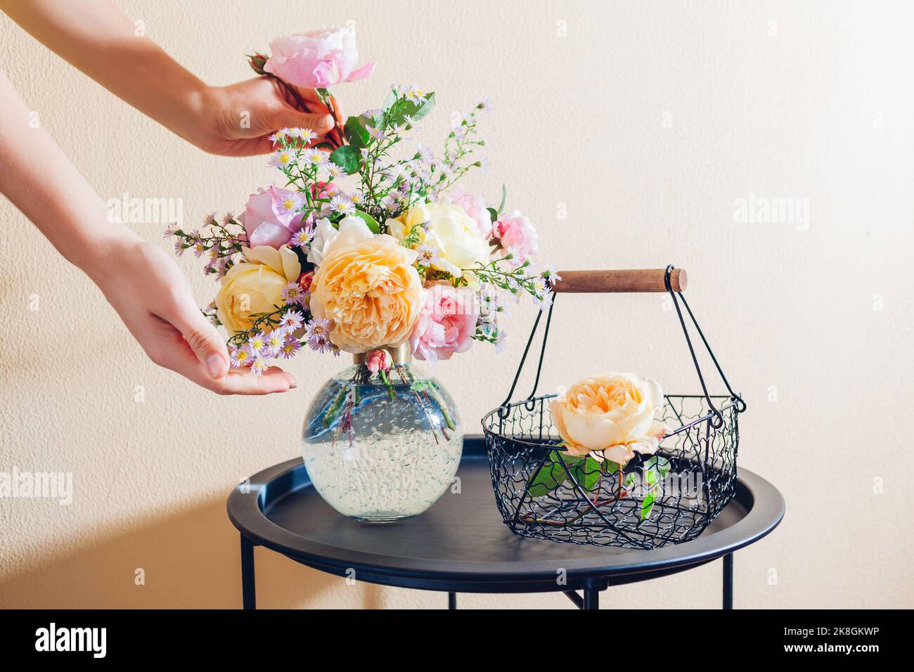 Frau macht Blumenstrauß in Vase aus orange gelb rosa Blumen auf dem Tisch zu Hause. Nahaufnahme von frischen englischen Rosen mit Metallkorb. Stockfoto