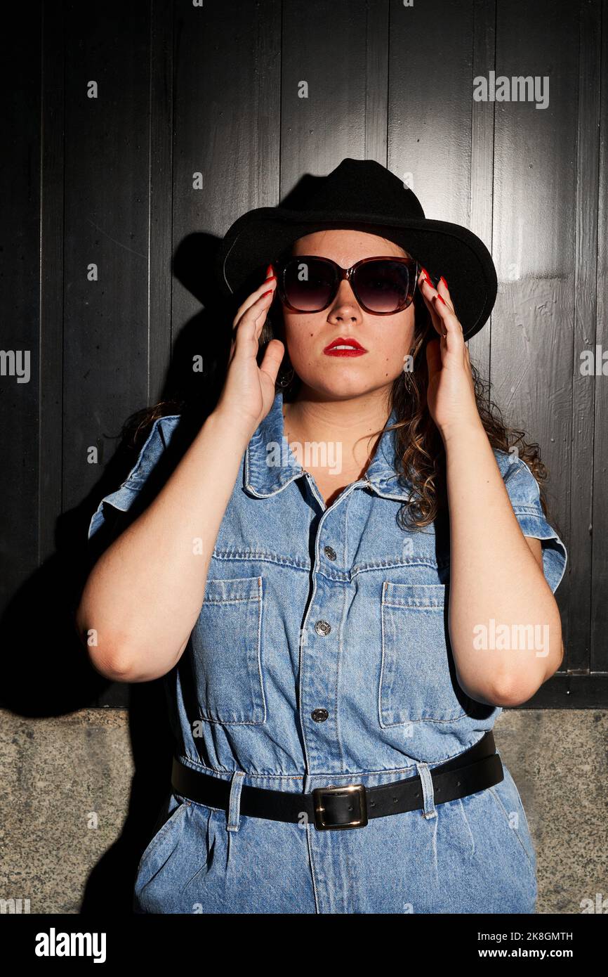 Junge Frau in Denim-Kleidung und stilvollem Hut, die Sonnenbrille anpasst und die Kamera gegen die schwarze Holzwand auf der Straße von Madrid, Spanien, anschaut Stockfoto