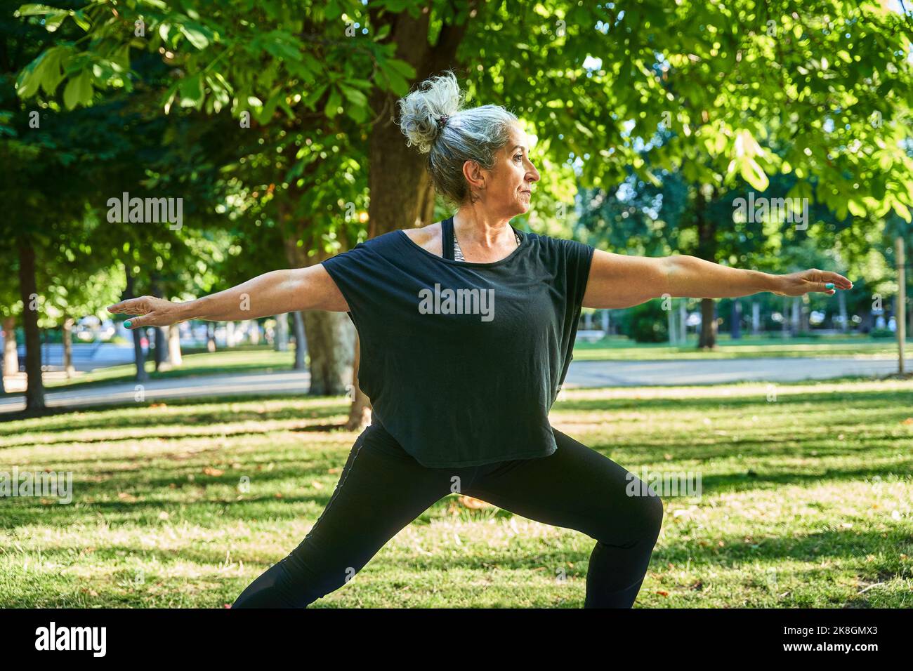Seitenansicht einer fokussierten älteren Frau in aktivem Ohr mit einem Bun, das in Virabhadrasana steht, während sie im Park unter grünen Blattbäumen auf der sonnigen da Yoga praktiziert Stockfoto