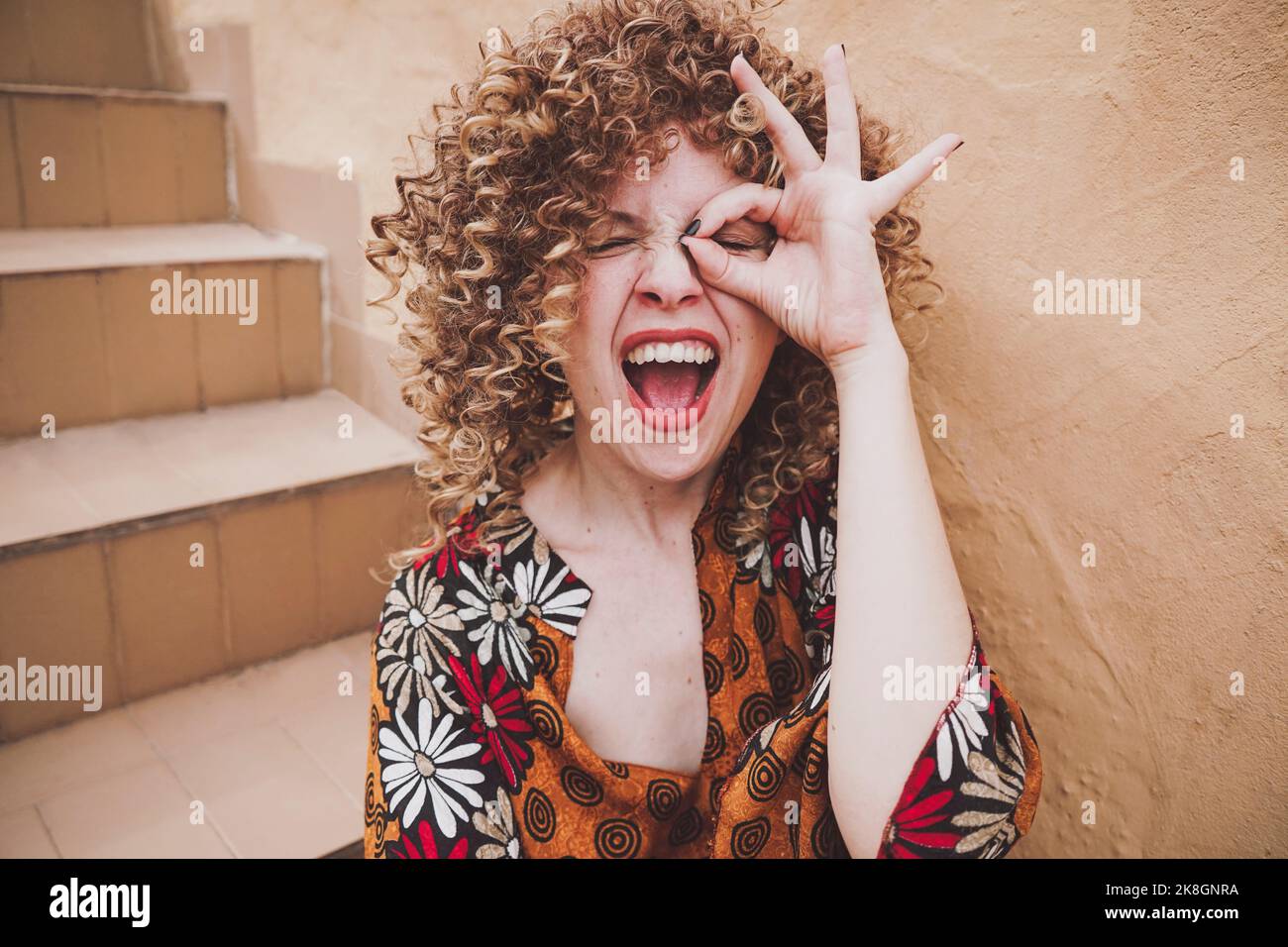 Entzückte junge Frau mit lockigen Haaren, die eine gute Geste nahe dem Auge an der Wand zeigte Stockfoto