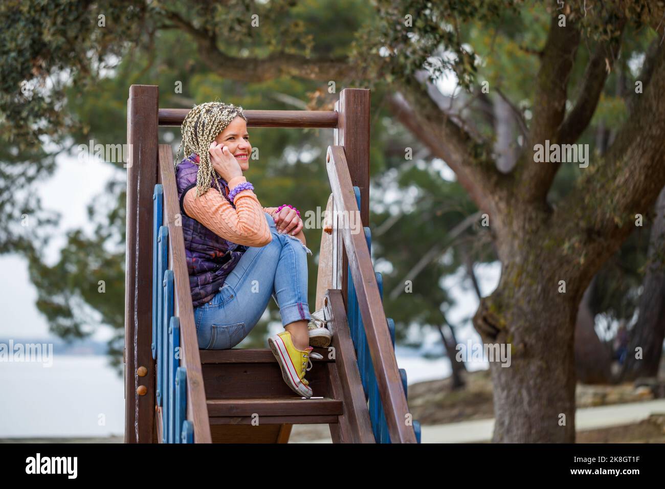 Attraktive reife Frau mit blonden Haaren und lässiger Kleidung, die in engem Raum auf der Rutsche auf dem Spielplatz mit dem Telefon sitzt Stockfoto