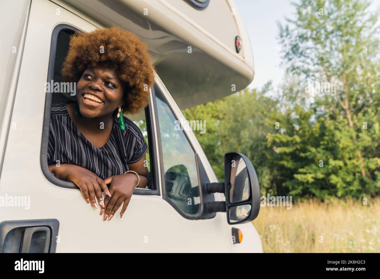 Schwarze Transporterfahrerin. Schöne positiv lächelnde schwarze Frau mit Afro-Frisur und langen Nägeln, die aus dem Fenster des Campers schauen. Reisekonzept. Außenaufnahme. Hochwertige Fotos Stockfoto