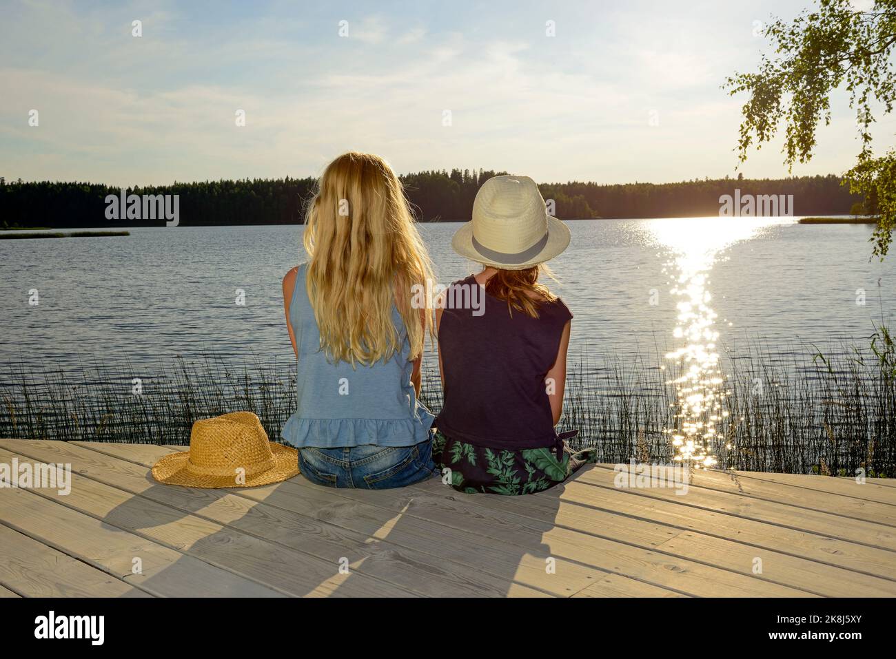 Zwei junge Mädchen von hinten sitzen auf einer Holzterrasse und blicken auf einen See bei Sonnenuntergang Stockfoto