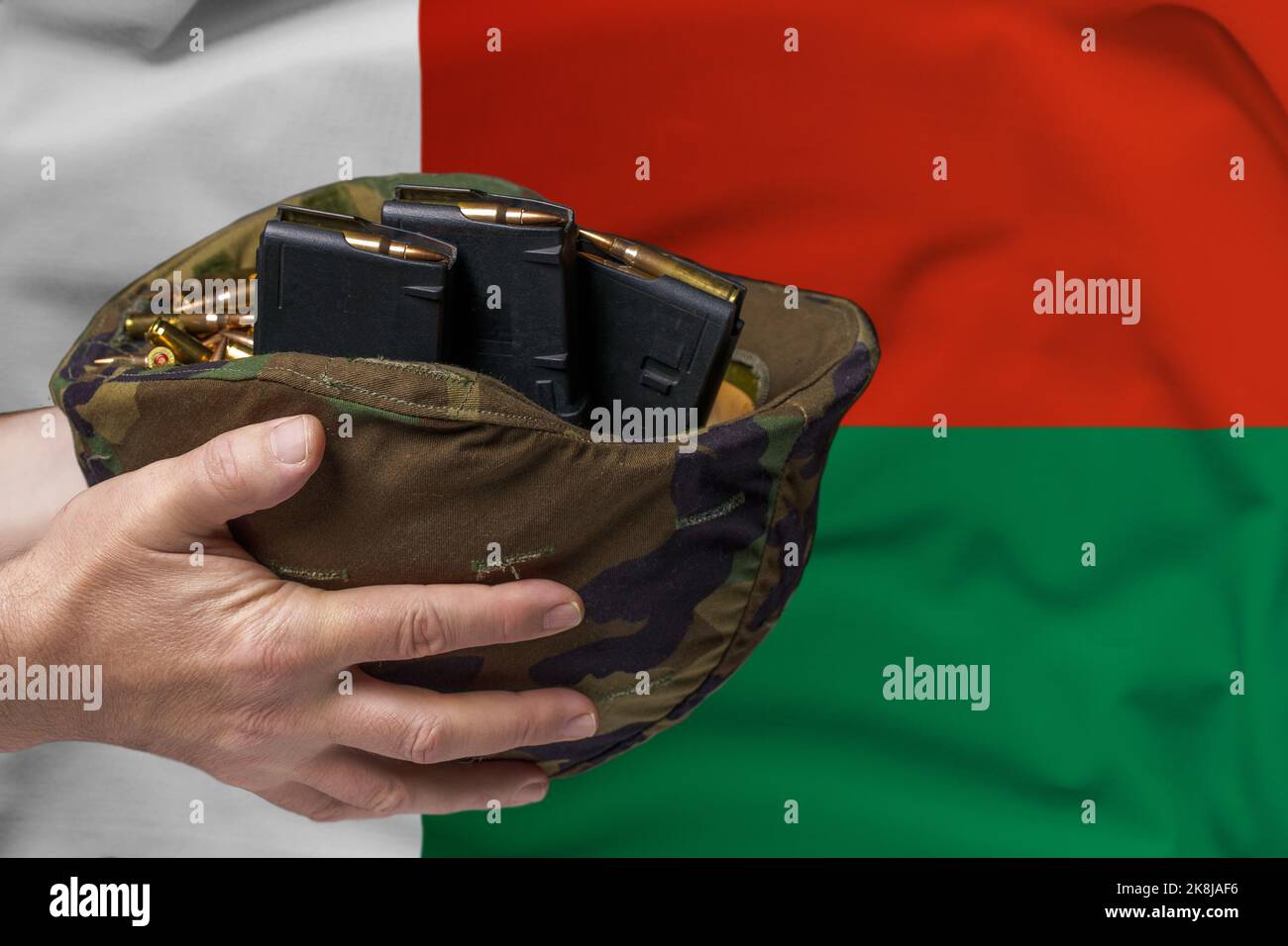 Ein Militärhelm mit Patronen und Zeitschriften für ein Gewehr in den Händen eines Mannes vor dem Hintergrund der Flagge Madagaskars. Das Konzept von Stockfoto