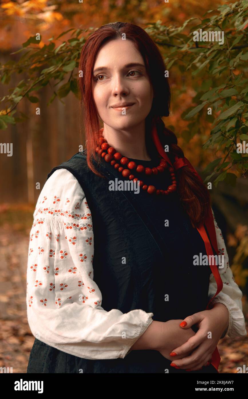 Lächelnde junge Rothaarige Frau, die traditionelle ukrainische bestickte Kleidung und rote Perlen angezogen hat, schaut weg. Vintage-Outfit. Halblanges Hochformat Stockfoto