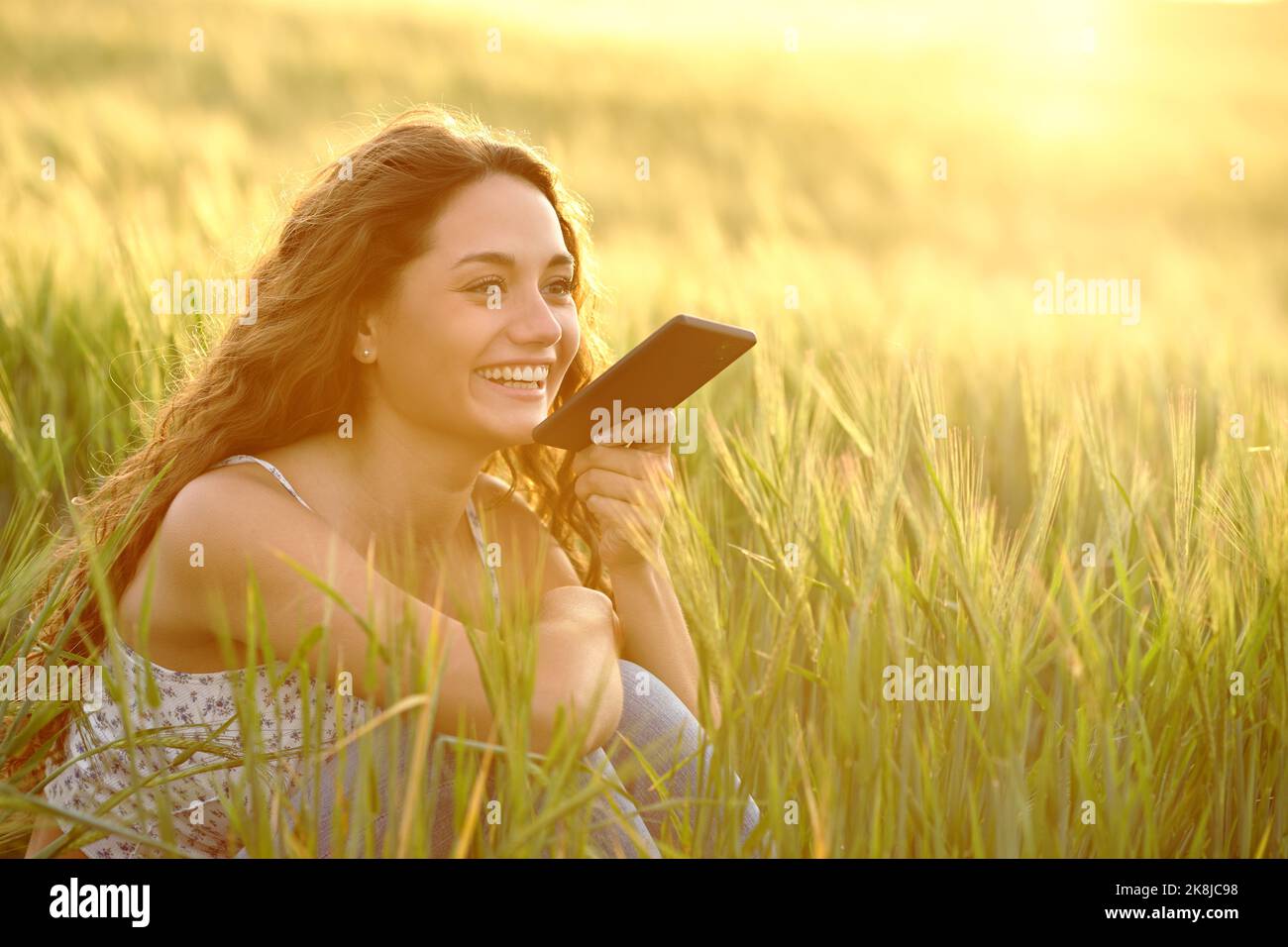Glückliche Frau, die bei Sonnenuntergang die Spracherkennung auf dem Smartphone in einem Weizenfeld verwendet Stockfoto