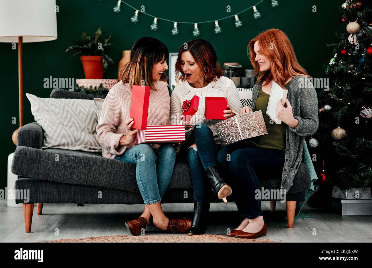 Drei attraktive Frauen mittleren Alters öffnen Geschenke zusammen, während sie während der Weihnachtszeit auf einem Sofa sitzen. Stockfoto