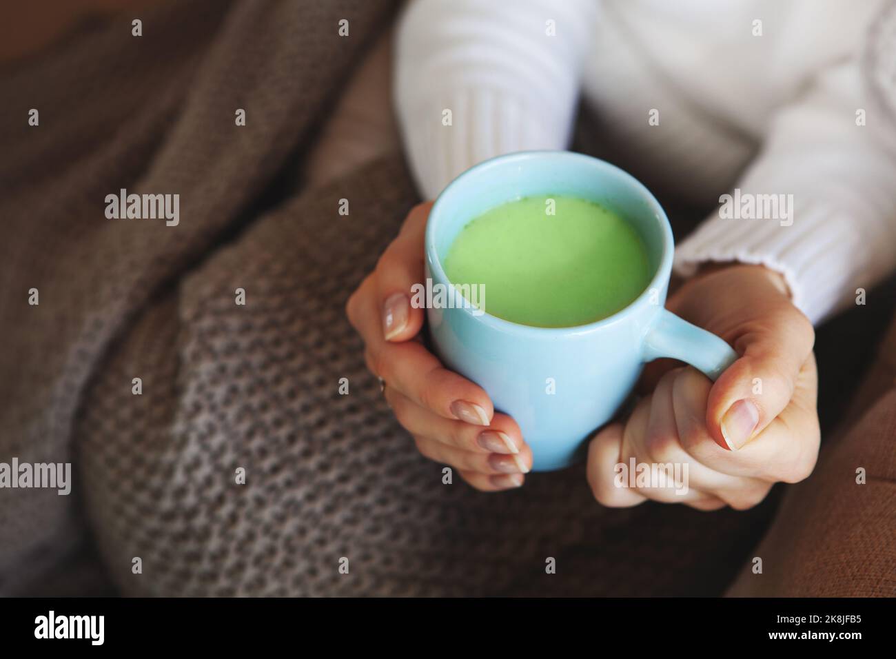 Nahaufnahme der Hände mit einer blauen Tasse Matcha-Tee. Modell in weißem Pullover und kuscheligem Karomell sitzt zu Hause. Schärfentiefe, leerer Raum auf verschwommener Beig Stockfoto