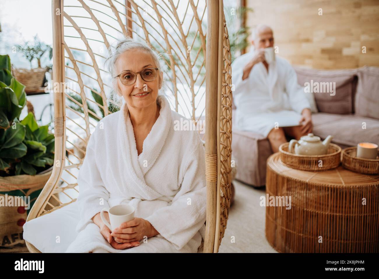 Ältere Frau, die auf einer Schaukel im Gebäude sitzt und die Kamera anschaut, ihr Mann genießt eine Tasse Tee im Hintergrund. Stockfoto