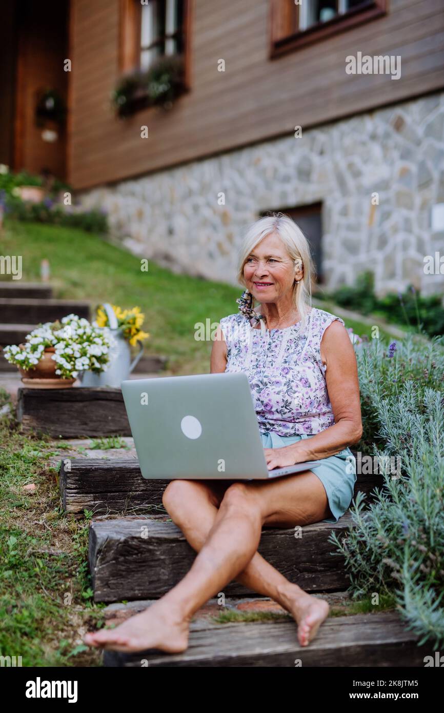 Ältere Frau, die Laptop verwendet und Bestellungen ihrer selbst angebauten Bio-Blumen und Gemüse im Garten, Konzept für kleine Unternehmen abwickeln. Stockfoto