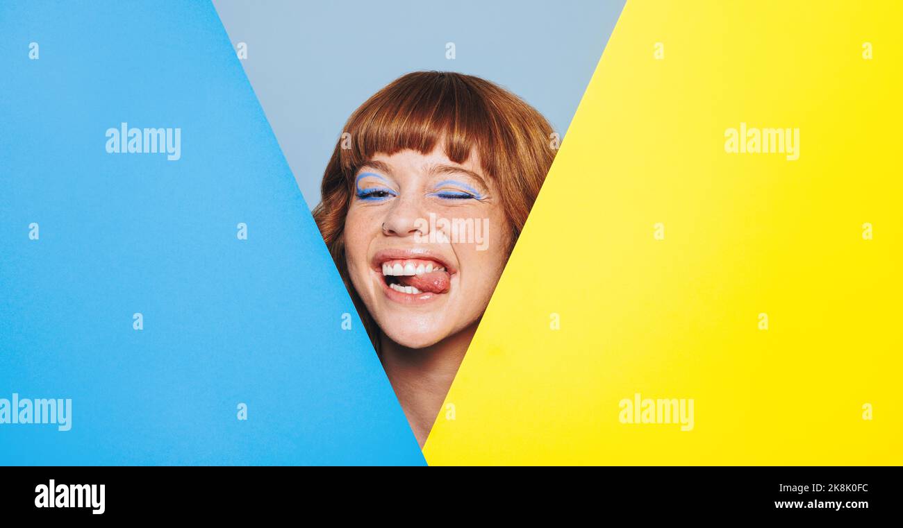 Schrullige Frau mit Ingwerhaar und blauem Lidschatten, die ihre Zunge herausstreckt, während sie hinter Farbplakaten steht. Verspielte junge Frau, die aus dem Hintern guckt Stockfoto