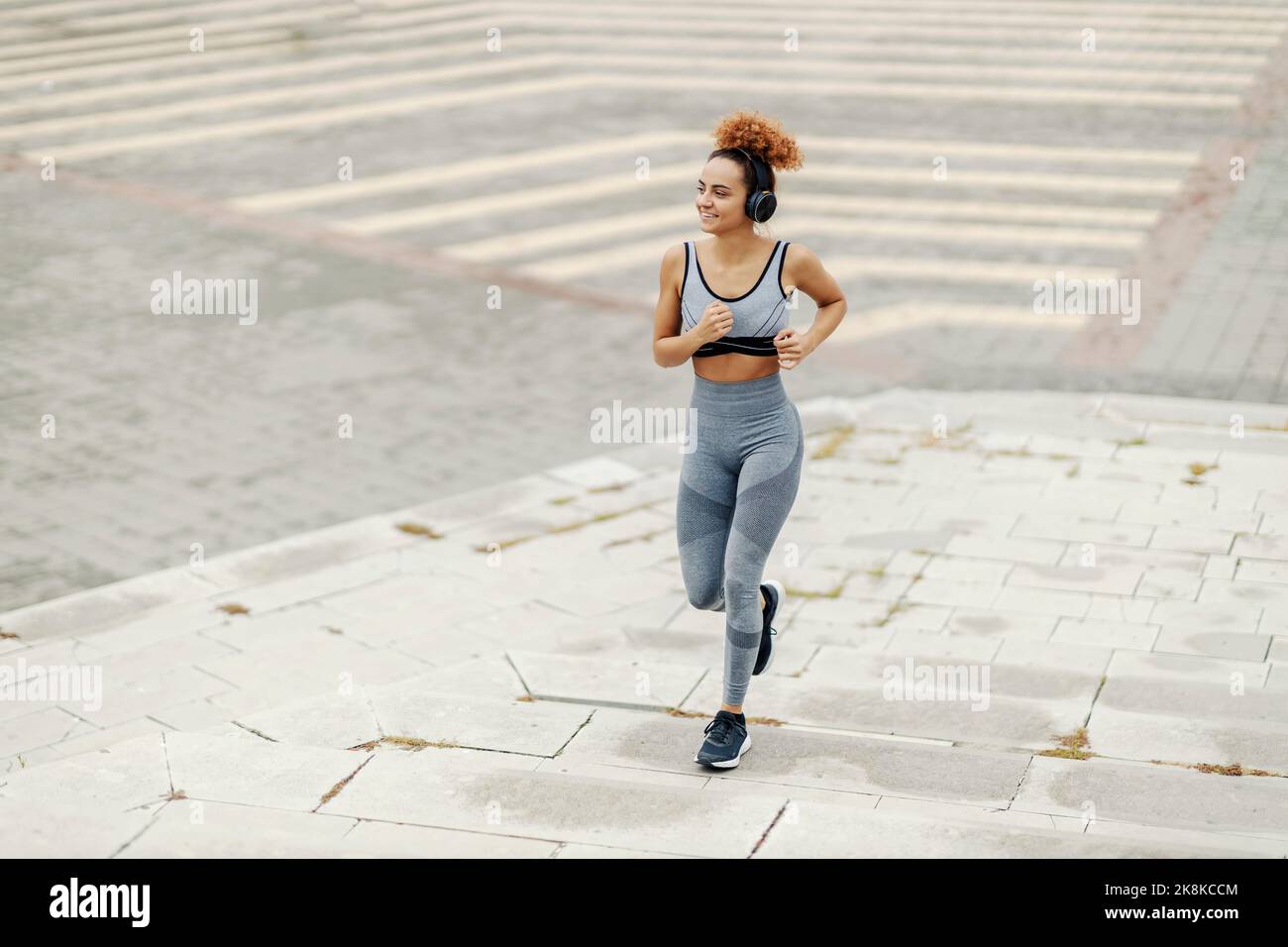 Die schnelle, fit gekleidete Sportlerin mit lockigen Haaren läuft mit Kopfhörern auf der Straße. Sie hört sich ihre Lieblingsradios an Stockfoto