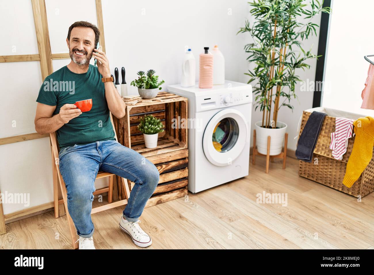 hispanischer Mann mittleren Alters, der Wäsche waschen und mit dem Smartphone reden und zu Hause Kaffee trinken. Stockfoto