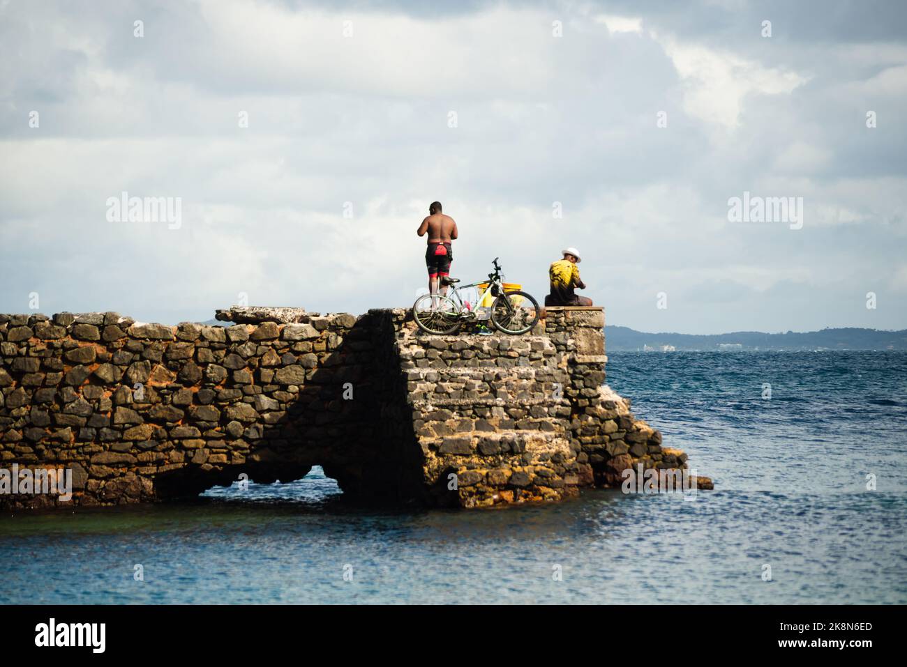 Die Menschen, die sich an sonnigen Tagen auf dem steinernen Wellenbrecher am Strand von Porto da Barra in Salvador ausruhen Stockfoto
